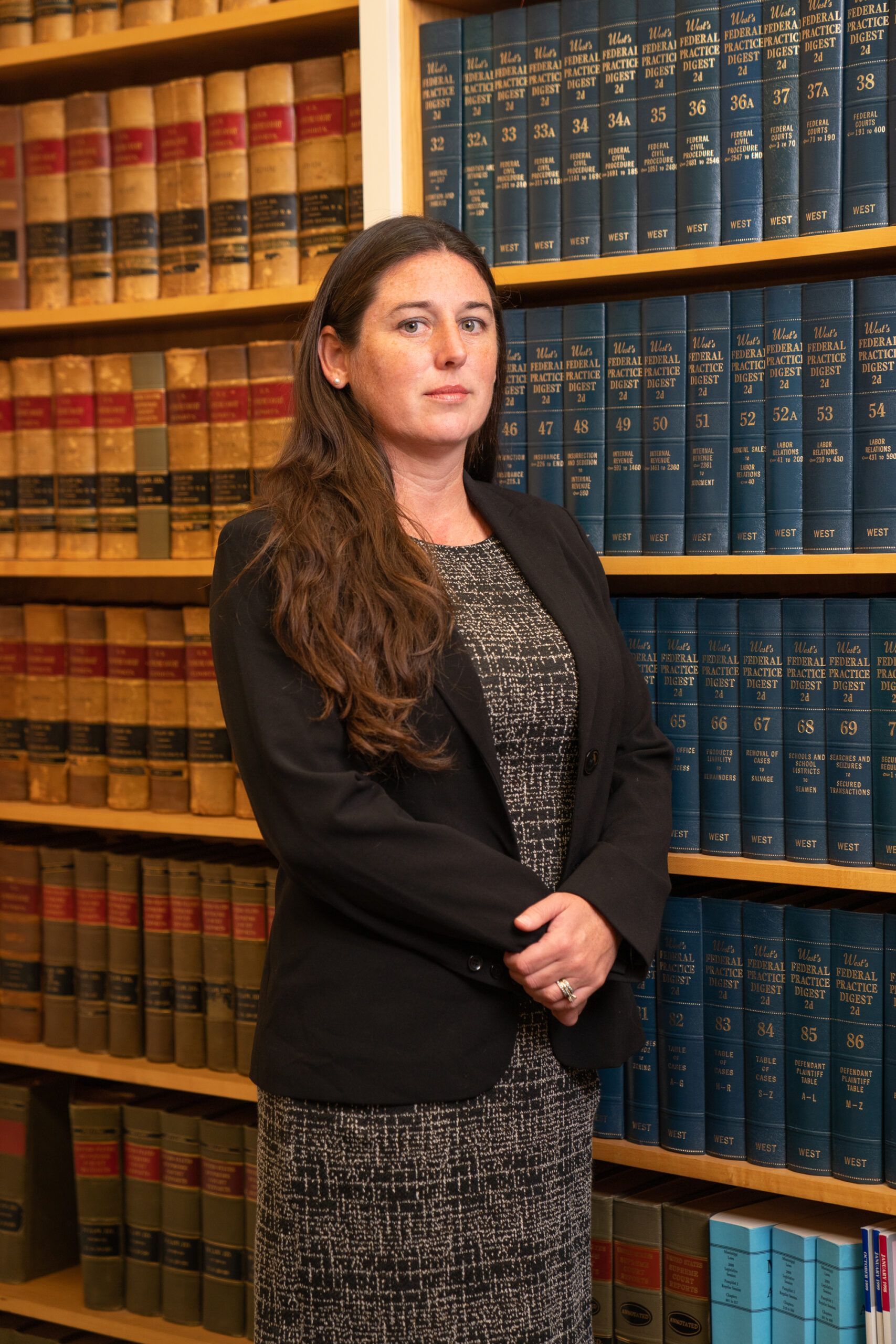 A person with long brown hair, wearing a black blazer and patterned dress, standing in front of a bookshelf full of books.