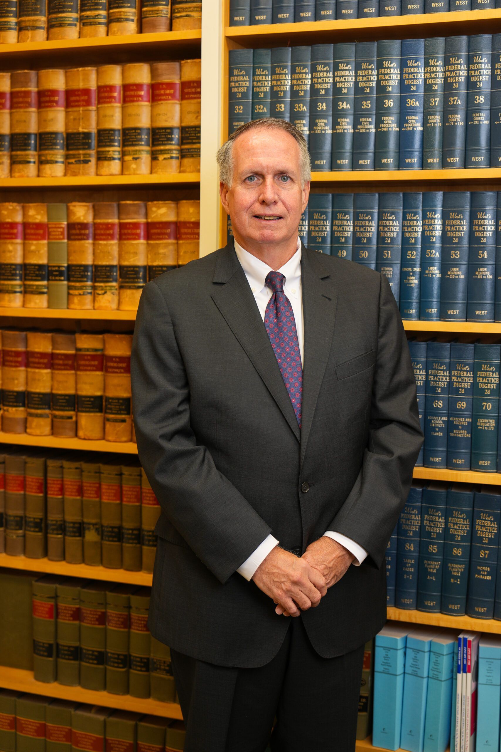 A professional in a suit and tie standing with hands clasped in front of a floor-to-ceiling bookshelf filled with law books.