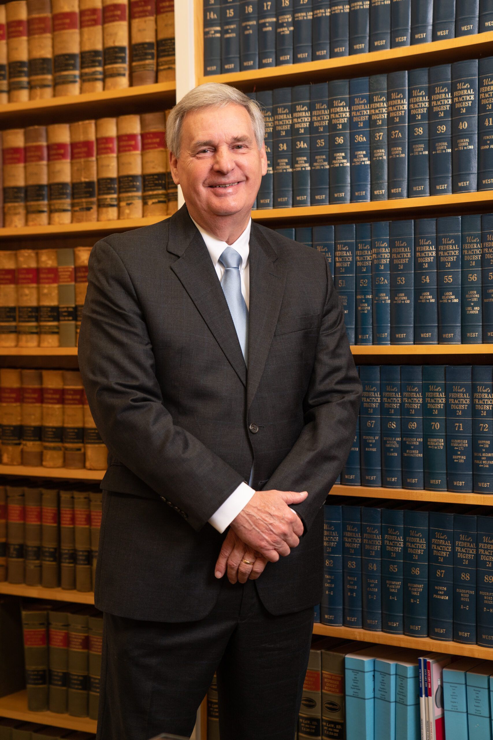 A man in a suit and light blue tie stands smiling in front of a bookshelf filled with many law books.