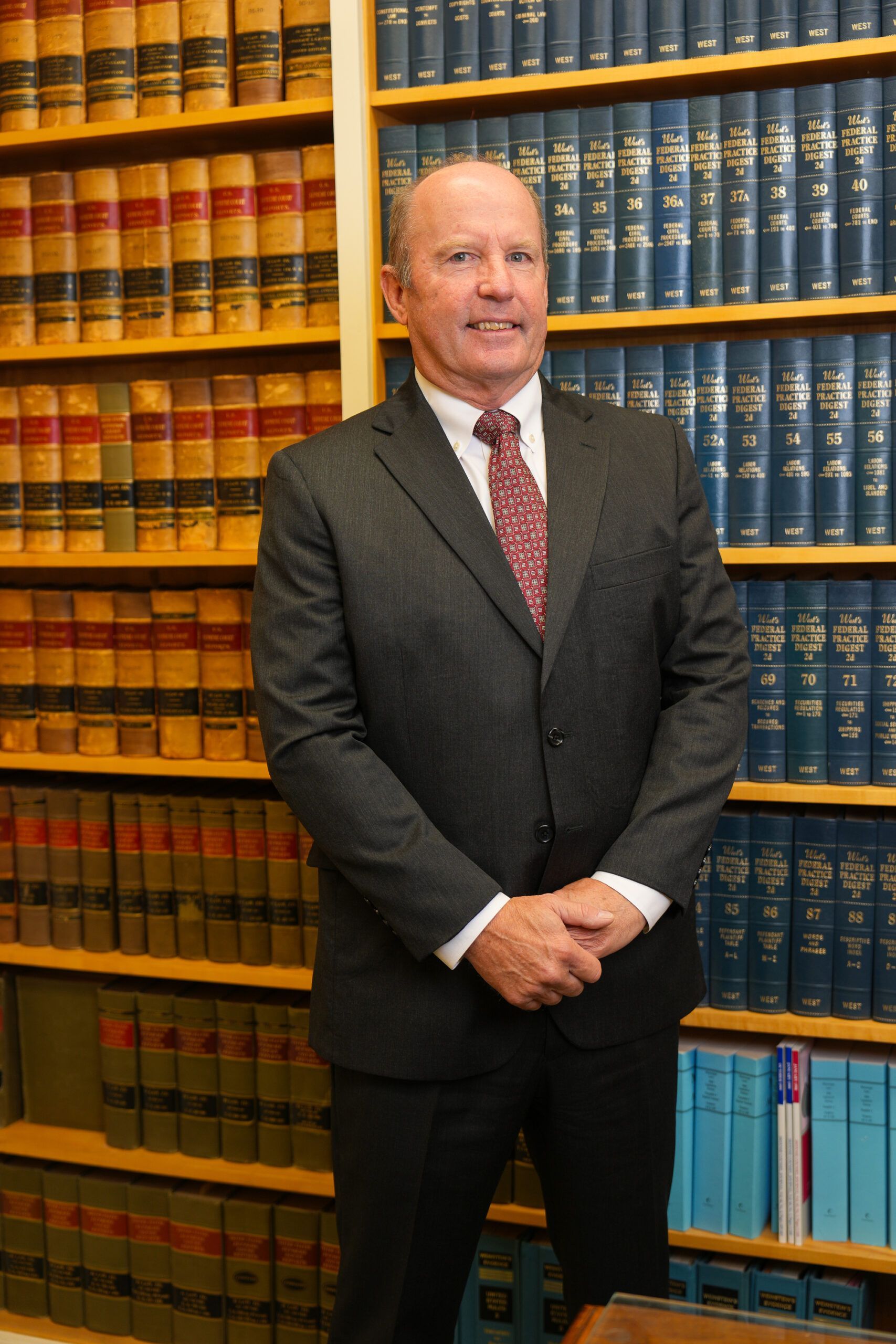 A person in a suit and patterned tie stands with hands clasped in front of shelves filled with law books.
