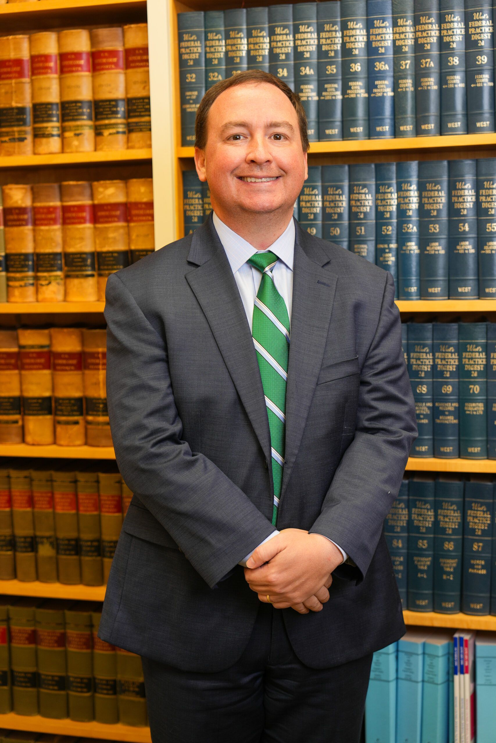 A person in a suit and green tie standing with hands clasped in front of a floor-to-ceiling bookshelf filled with books.