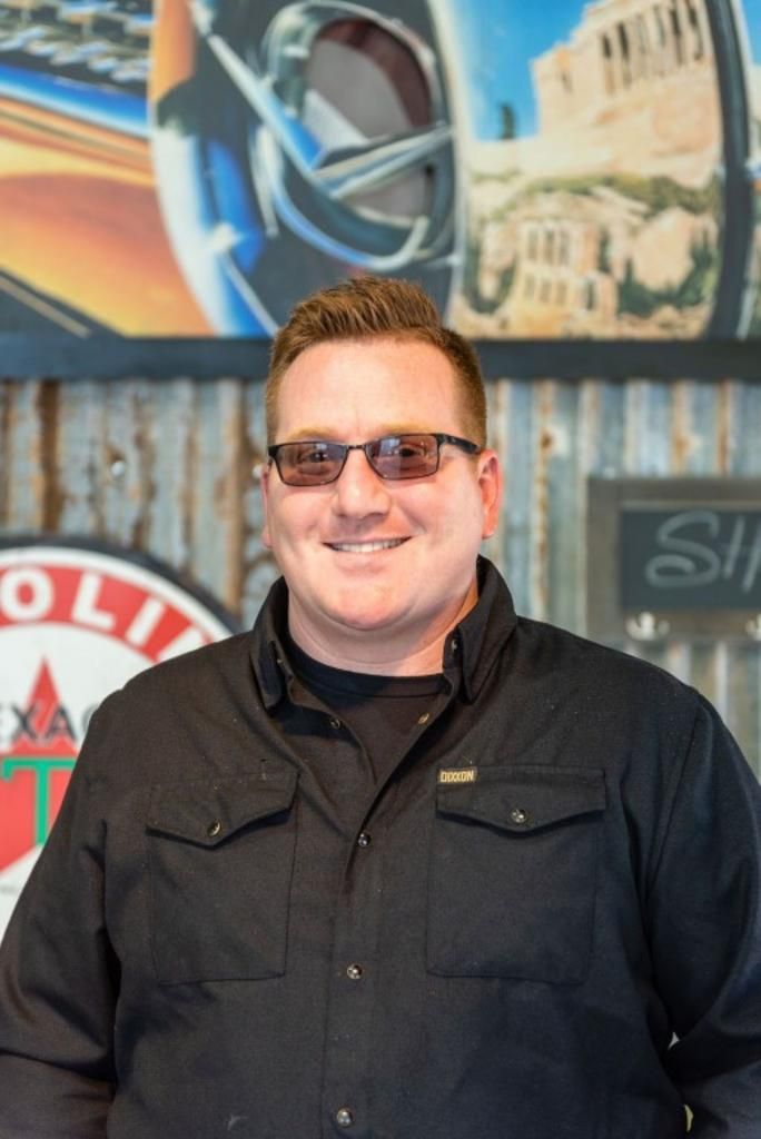 A man wearing sunglasses and a black jacket is smiling for the camera.