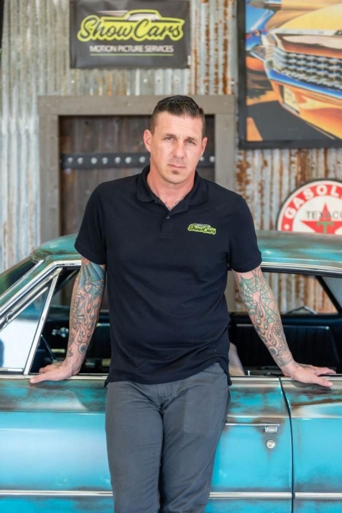 A man with tattoos is leaning against a blue car in a garage.