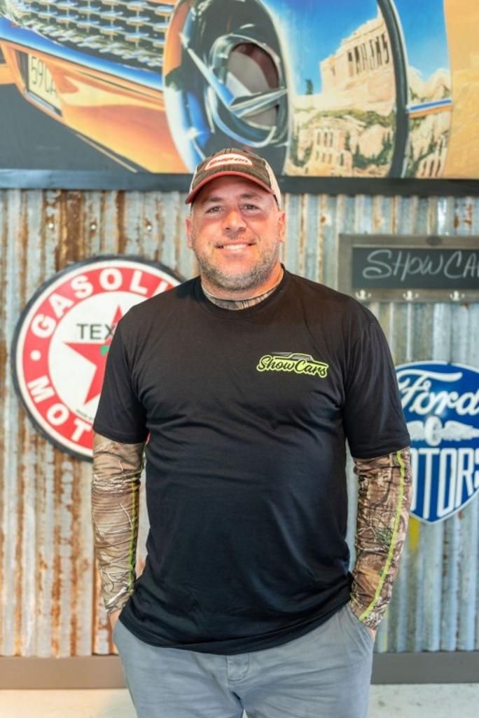 A man wearing a black t-shirt and a hat is standing in front of a metal wall.