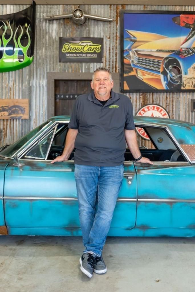 A man is leaning against a blue car in a garage.