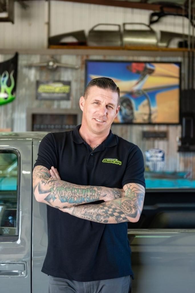 A man with tattoos on his arms is standing in front of a truck in a garage.