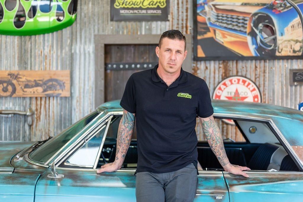 A man is leaning against a blue car in a garage.
