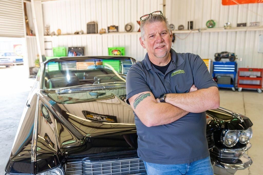 A man is standing in front of a black car with his arms crossed.