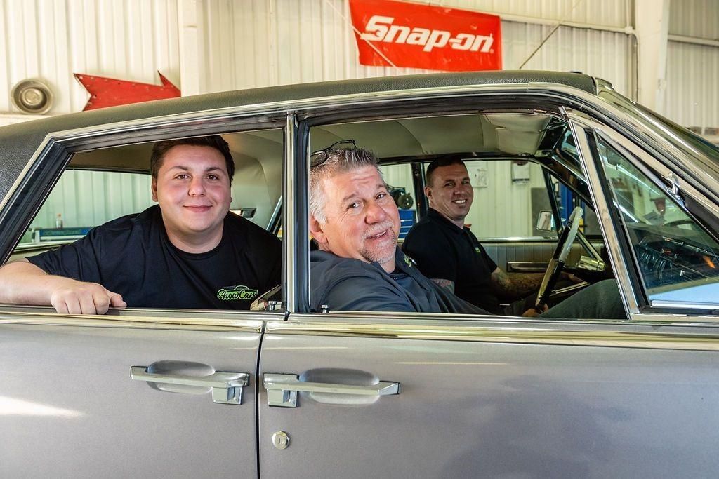 Three men are sitting in a car with a snap-on sign in the background.