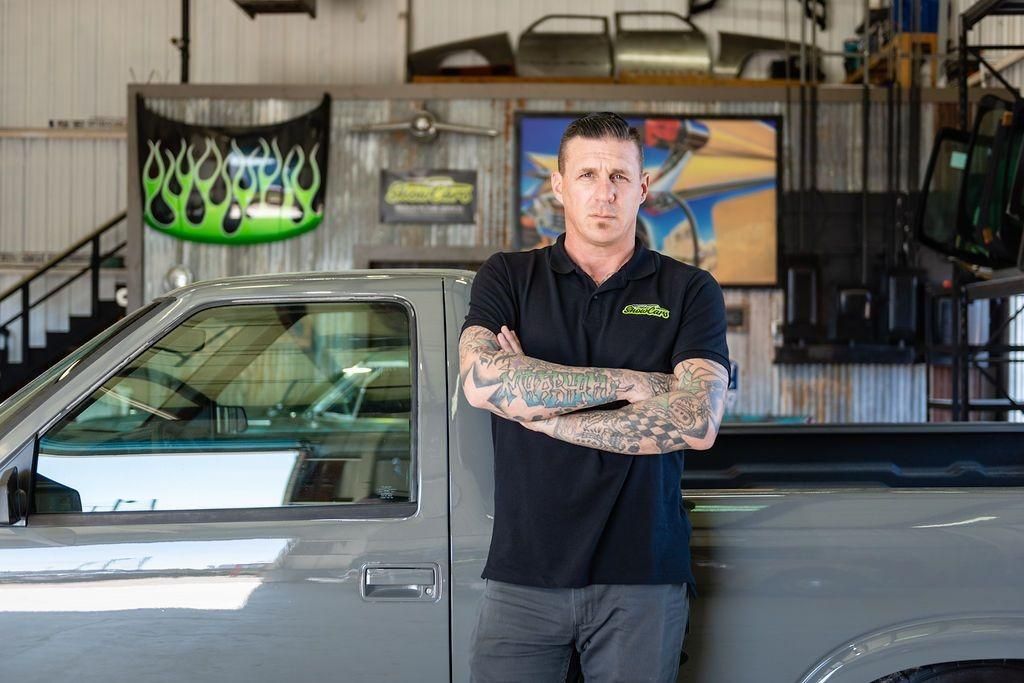 A man with tattoos is standing next to a truck in a garage.