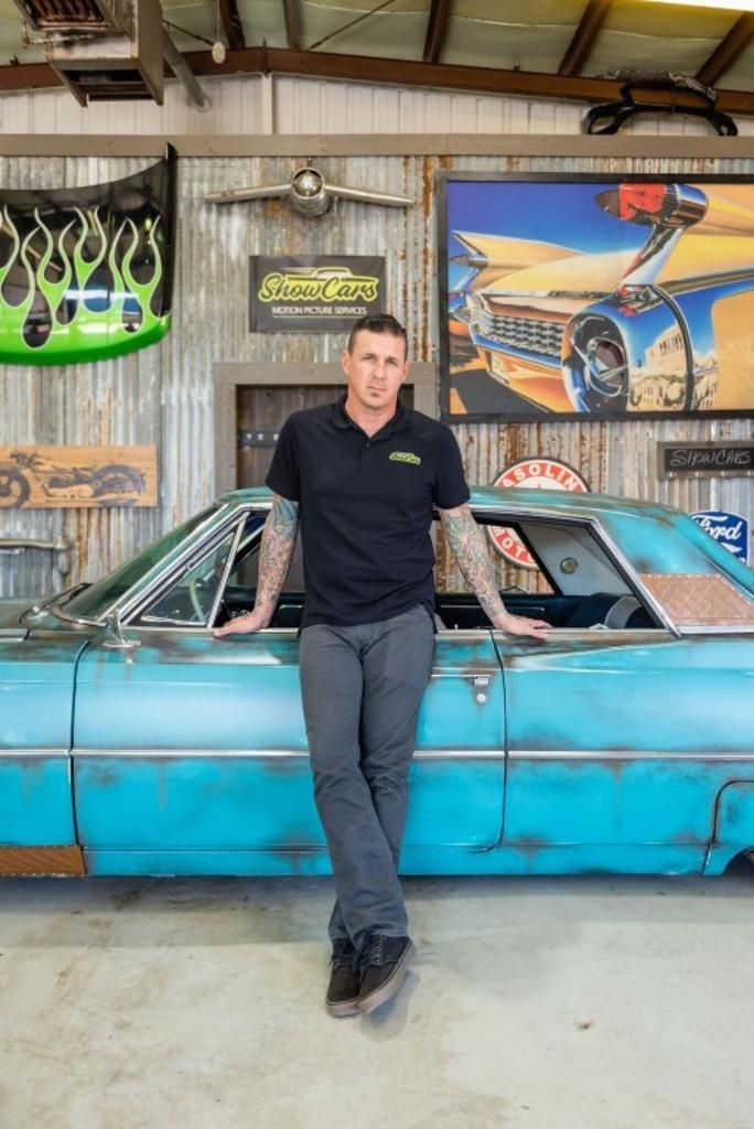 A man is leaning against a blue car in a garage.