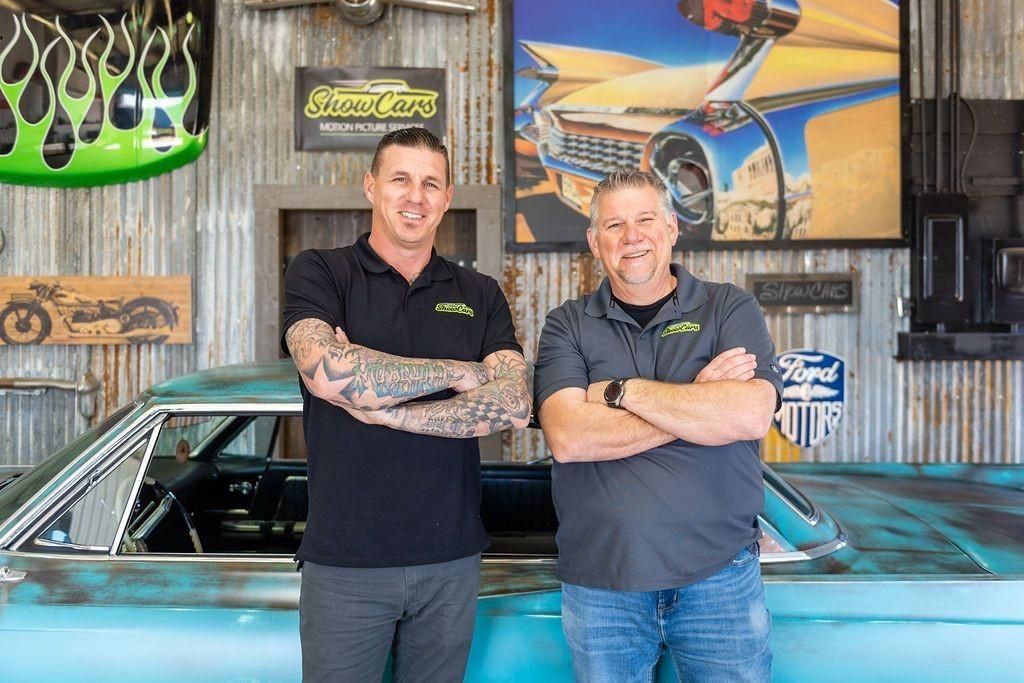 Two men are standing next to each other in front of a car in a garage.