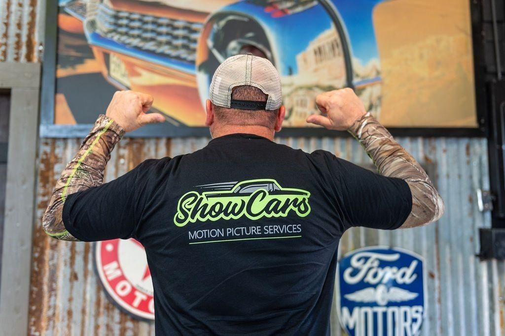 A man wearing a show cars t-shirt is flexing his muscles in front of a ford sign.