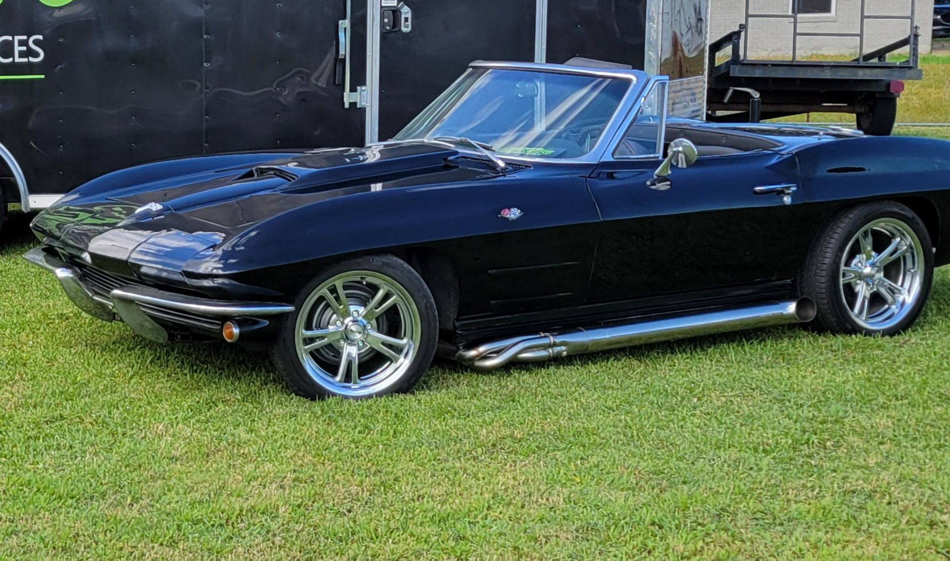 A black corvette convertible is parked in the grass in front of a trailer.