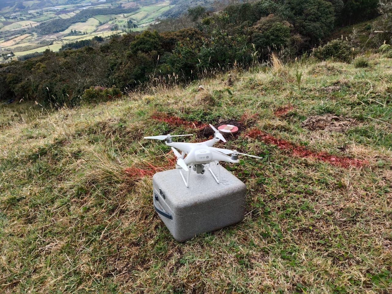 Dron blanco sobre una caja gris, en una ladera herbosa, con marcas rojas. Paisaje montañoso al fondo.
