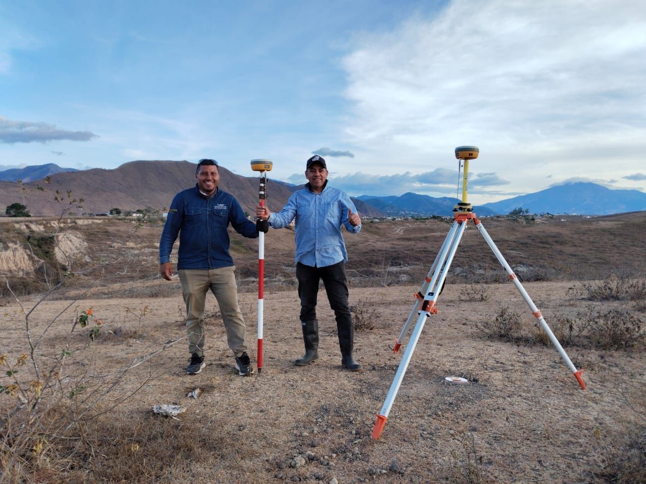 Dos hombres con equipo topográfico al aire libre; uno sosteniendo una vara, el otro junto a un trípode con un receptor GPS, montañas al fondo.