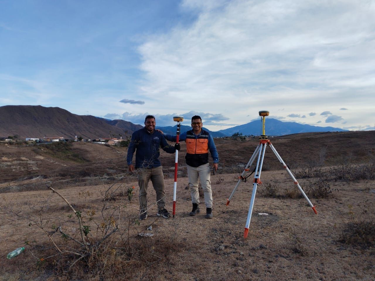 Dos hombres con equipo topográfico al aire libre; campo, colinas y cielo visibles.