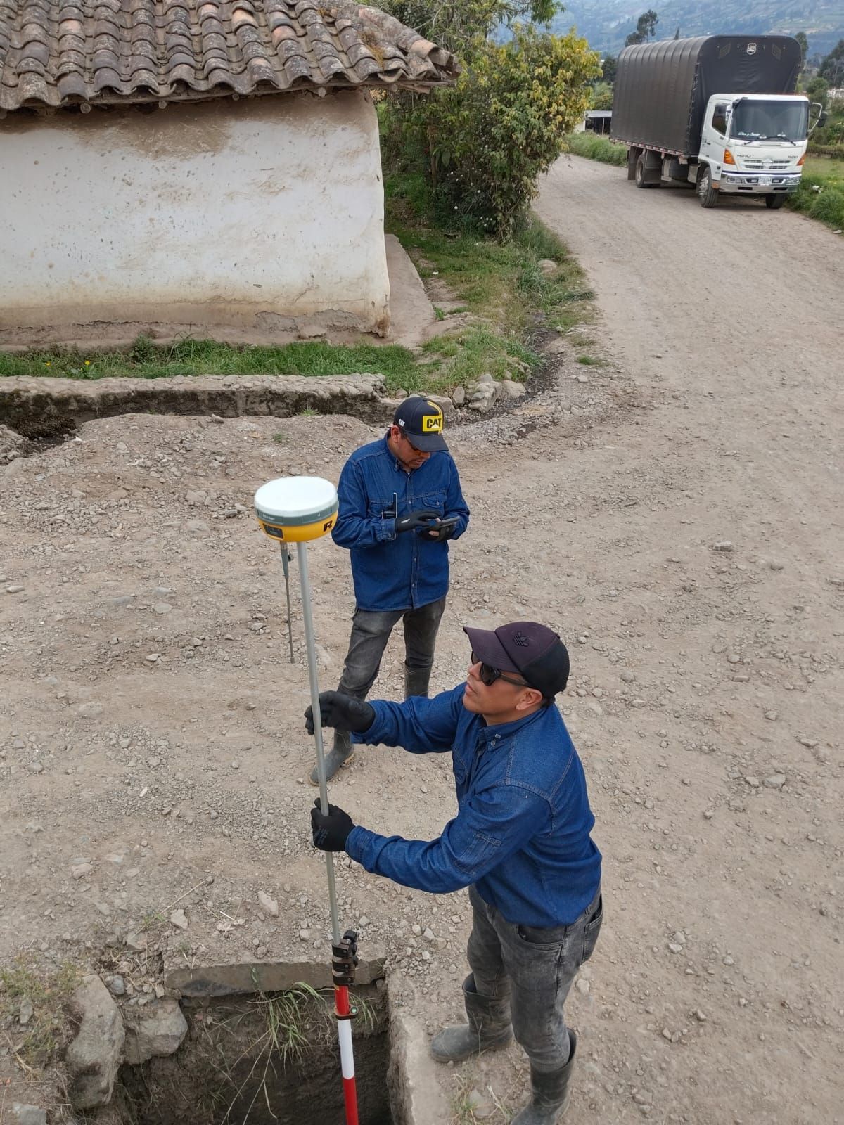 Dos hombres utilizan equipo de topografía en un camino de tierra cerca de un pequeño edificio; camión al fondo.