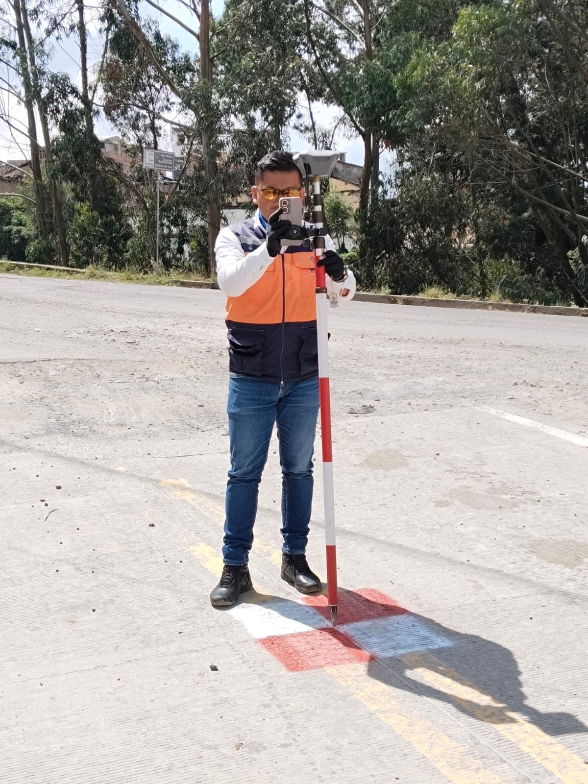Hombre sosteniendo un equipo de topografía en una carretera, de pie cerca de un marcador rojo y blanco.