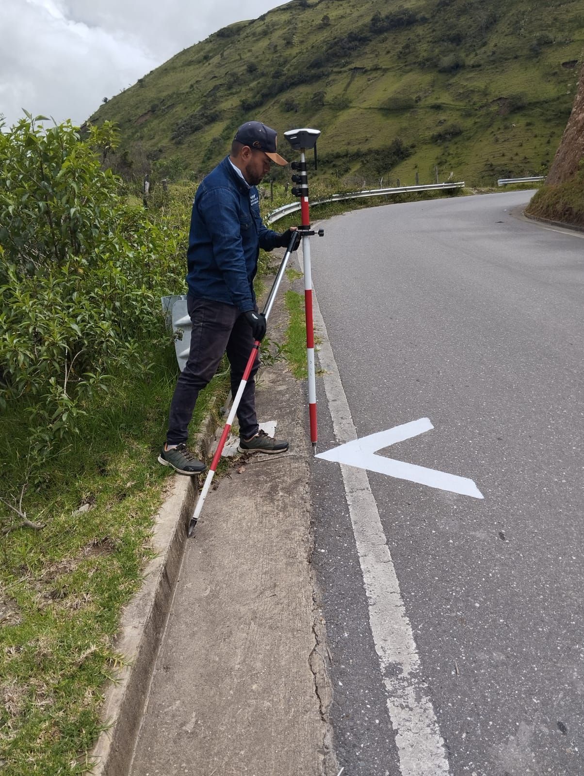 Hombre inspeccionando la carretera con equipo GPS, de pie cerca de una flecha pintada en el asfalto al lado de la acera