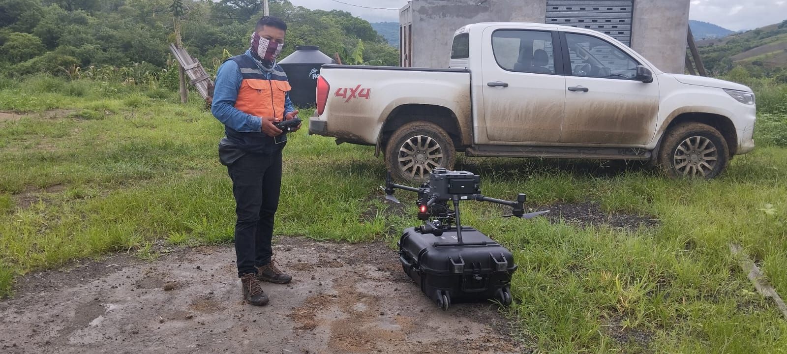 Una persona con un chaleco naranja opera un dron negro cerca de una camioneta blanca en un campo de hierba.