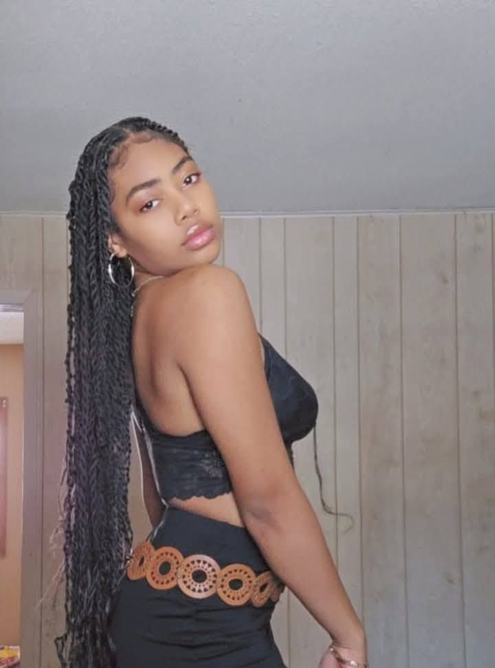 A woman with long braids is wearing a black top and a brown belt.