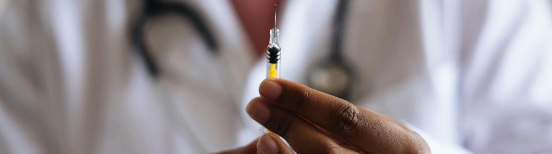 Person with dark skin holding a vaccine syringe. They are wearing a white coat,