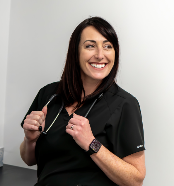 Smiling doctor in a white coat holds a tablet, hospital hallway with other medical staff in the background.