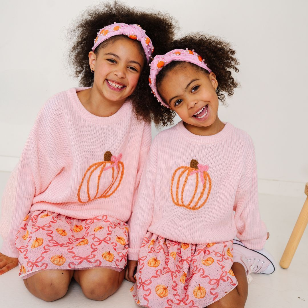 Two smiling girls in matching pink sweaters and skirts, each with a pumpkin design, wearing pumpkin headbands.