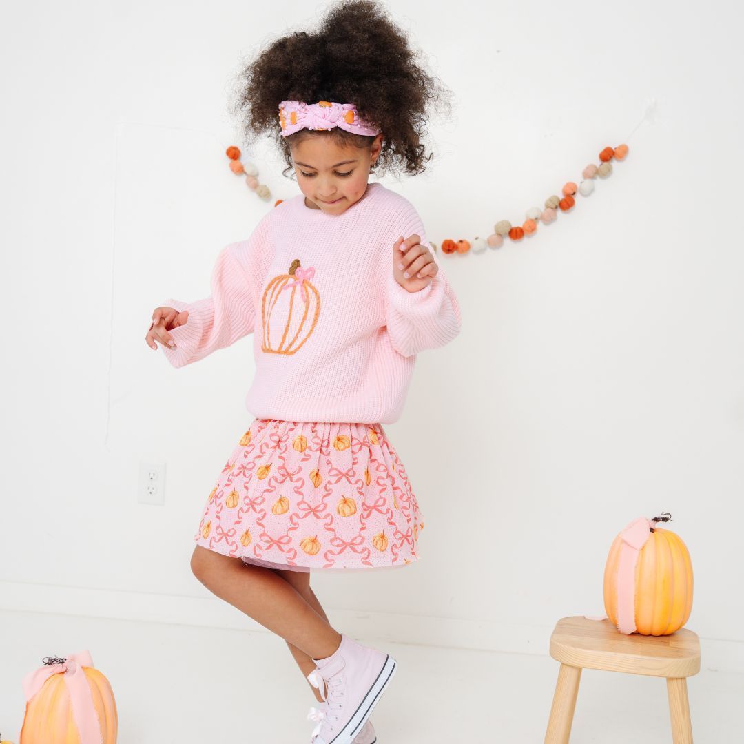 Young girl in a pink pumpkin-themed outfit jumping. She wears a sweater, skirt, and headband. A pumpkin sits near a stool.