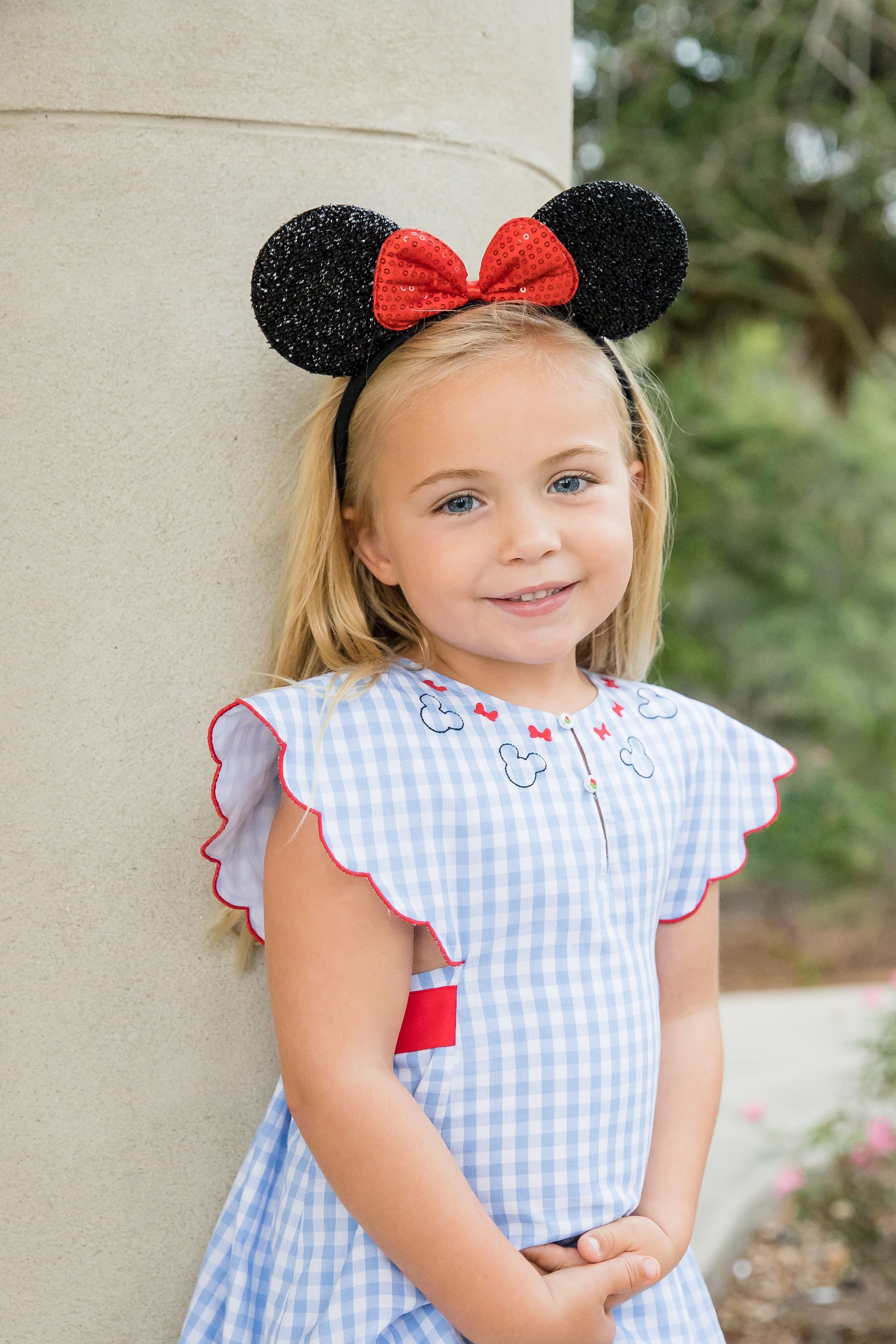 A little girl wearing a minnie mouse headband is standing next to a wall.