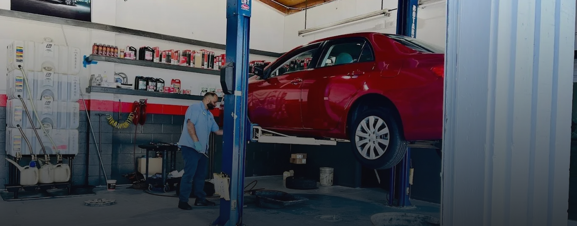 A red car is elevated on a lift in an auto repair shop; a mechanic works on the car near shelves with supplies.