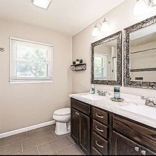 Modern beige bathroom with double mirrors, dark vanity, toilet, and window.