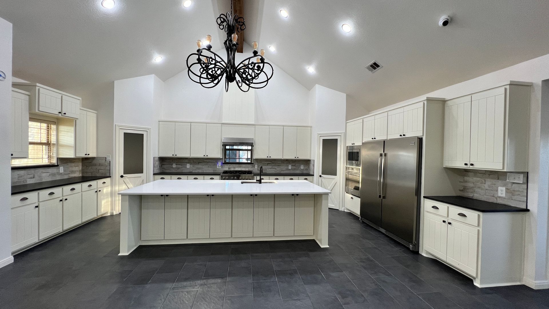 Spacious white kitchen with a large island, stainless steel fridge, black chandelier, and dark tile floor.