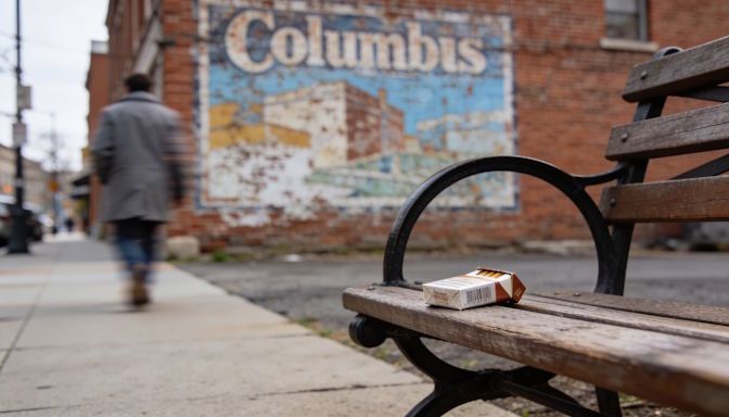Short North street scene, discarded cigarette pack resting on bench, lone figure walking away, blurred Columbus mural