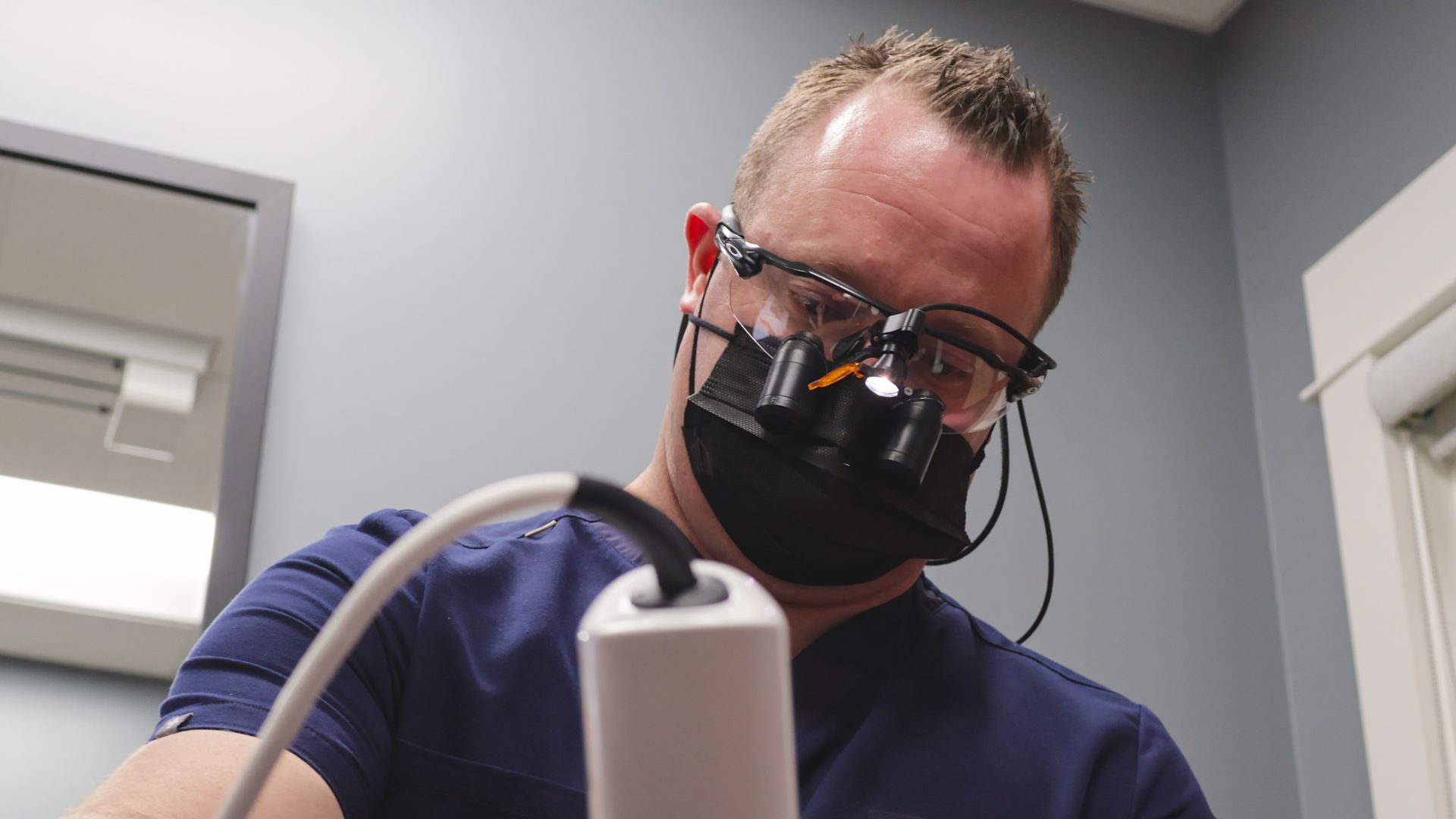 Dentist and assistant treating a patient in a dental office. Both wear masks, patient reclined.