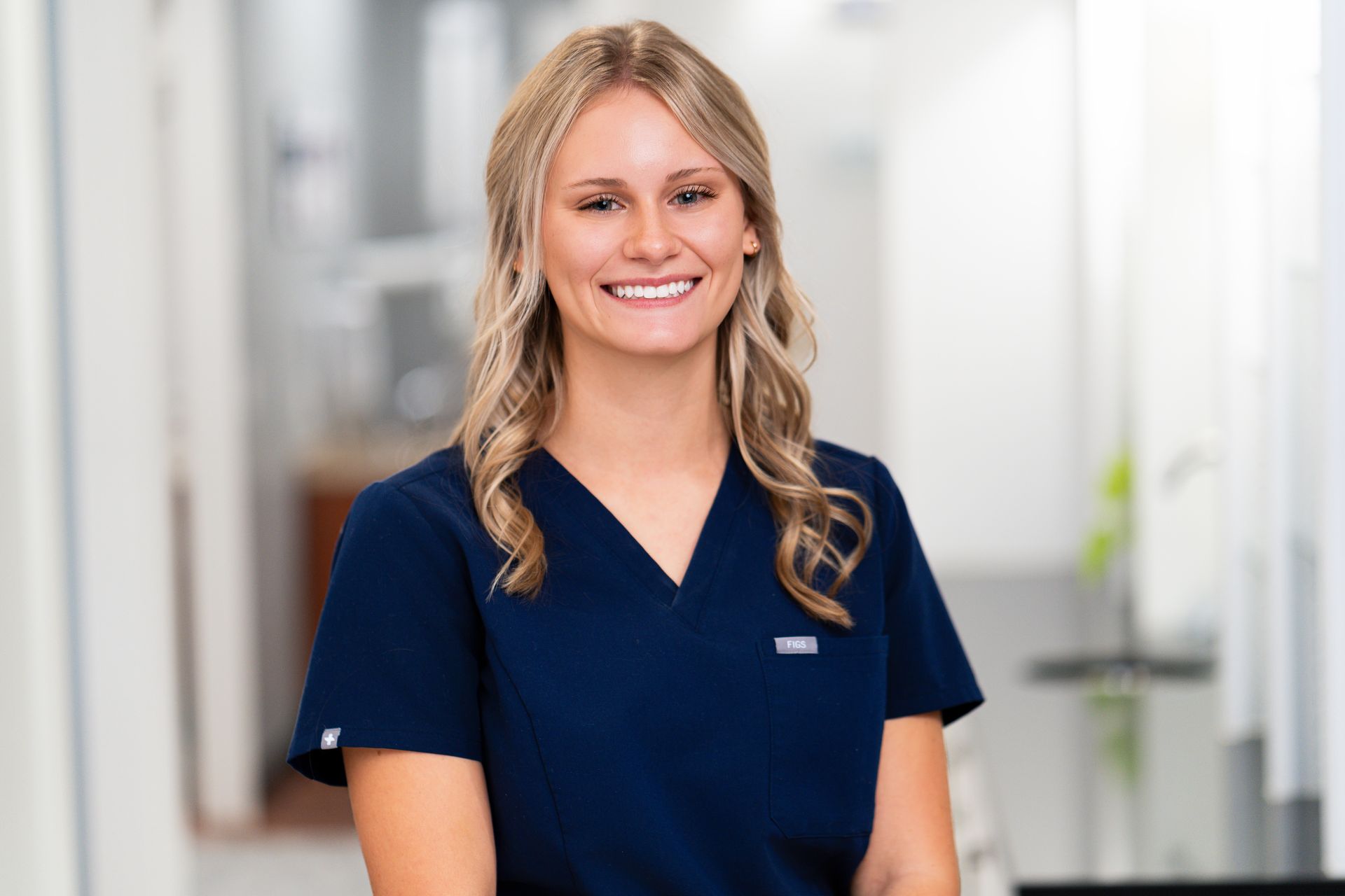 Woman in dark blue scrubs smiles in a brightly lit hallway.