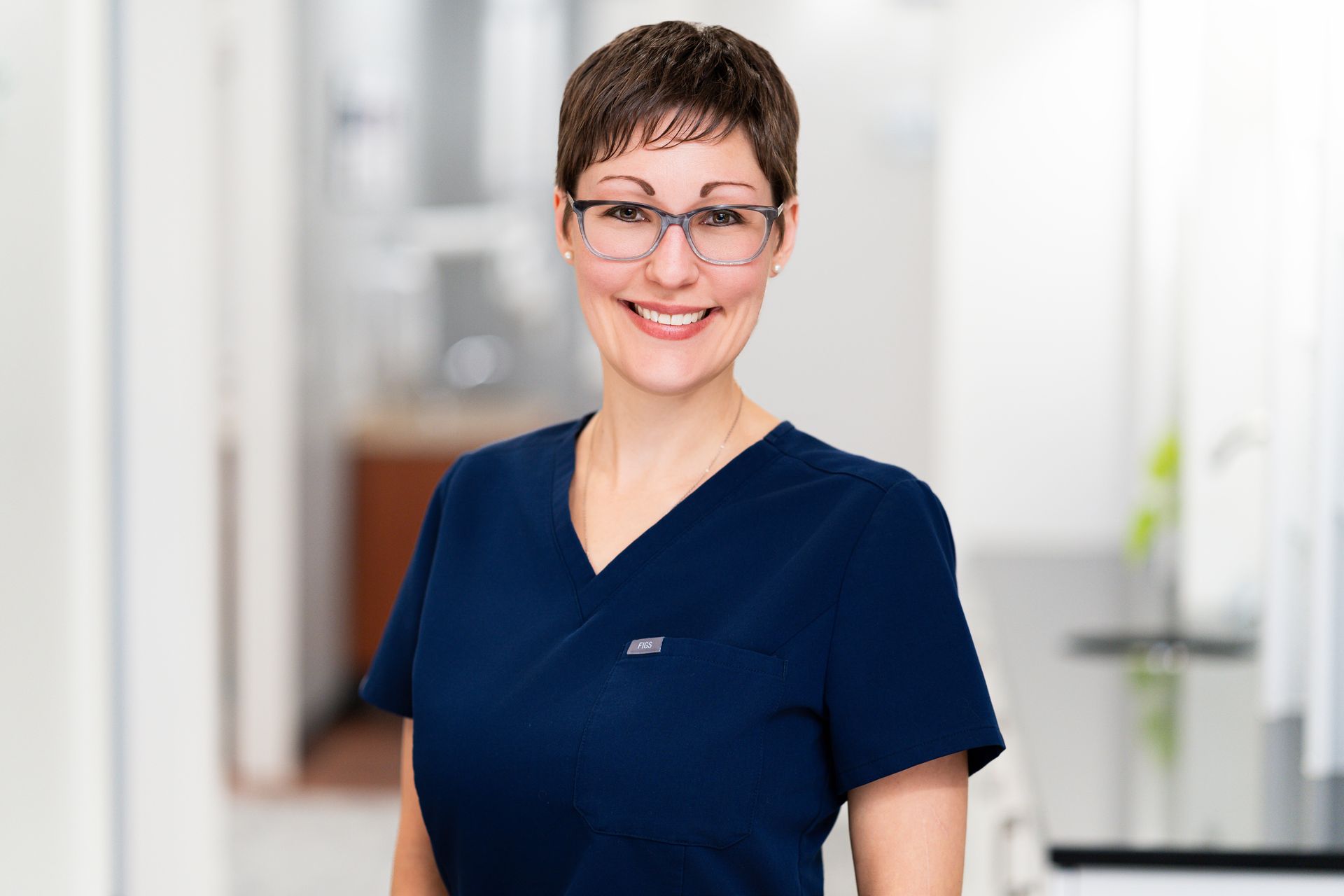 Woman in black scrubs smiles at the camera, standing by a white desk in a dental office.