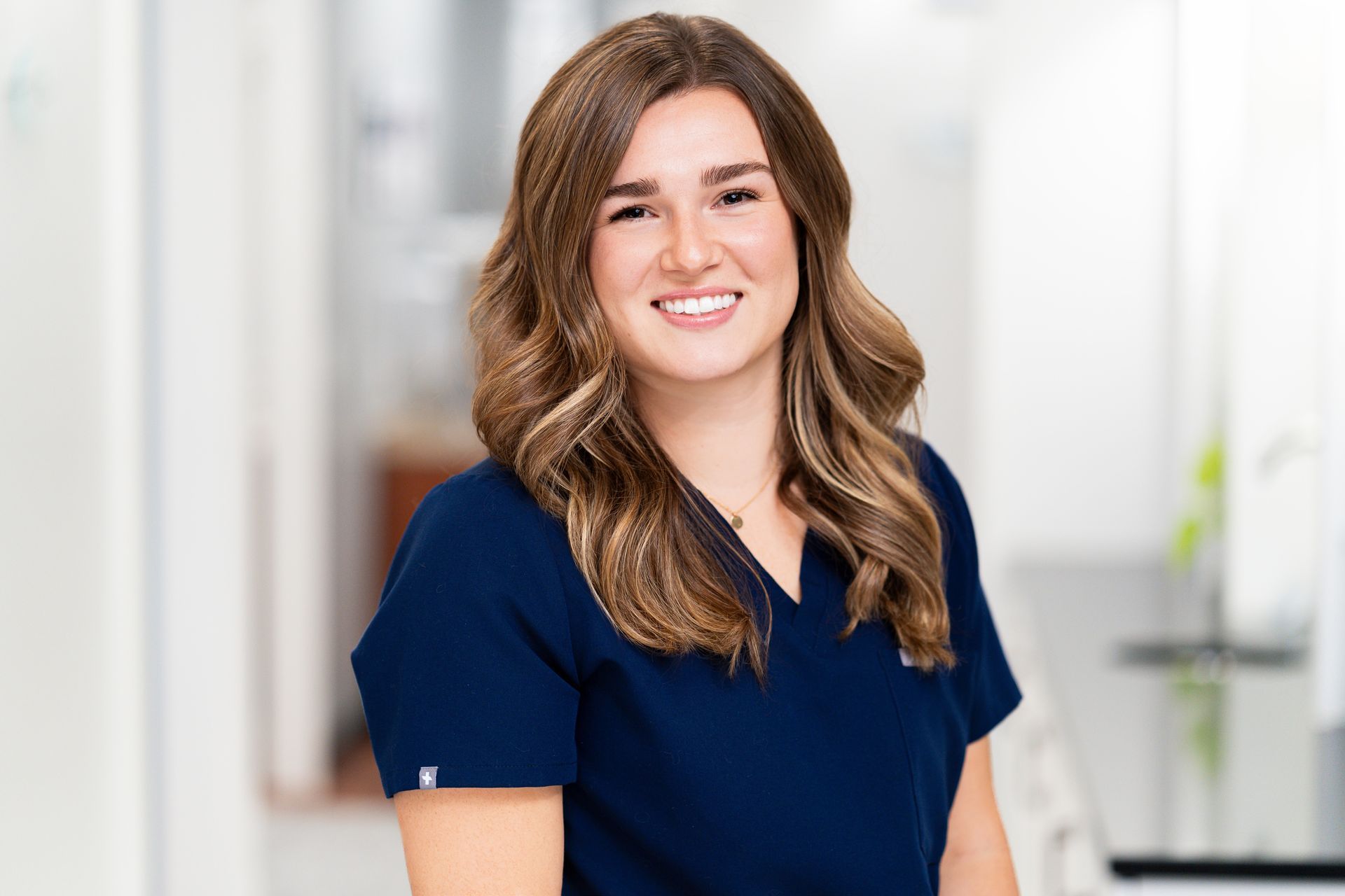 Woman with brown wavy hair smiles in a blue uniform, likely in a medical setting.