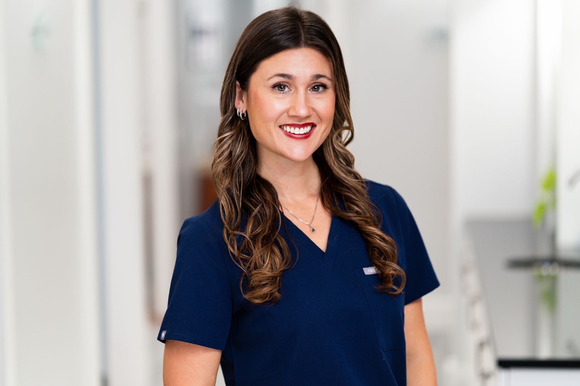 Woman with long brown hair, wearing navy scrubs, smiling in a well-lit office hallway.