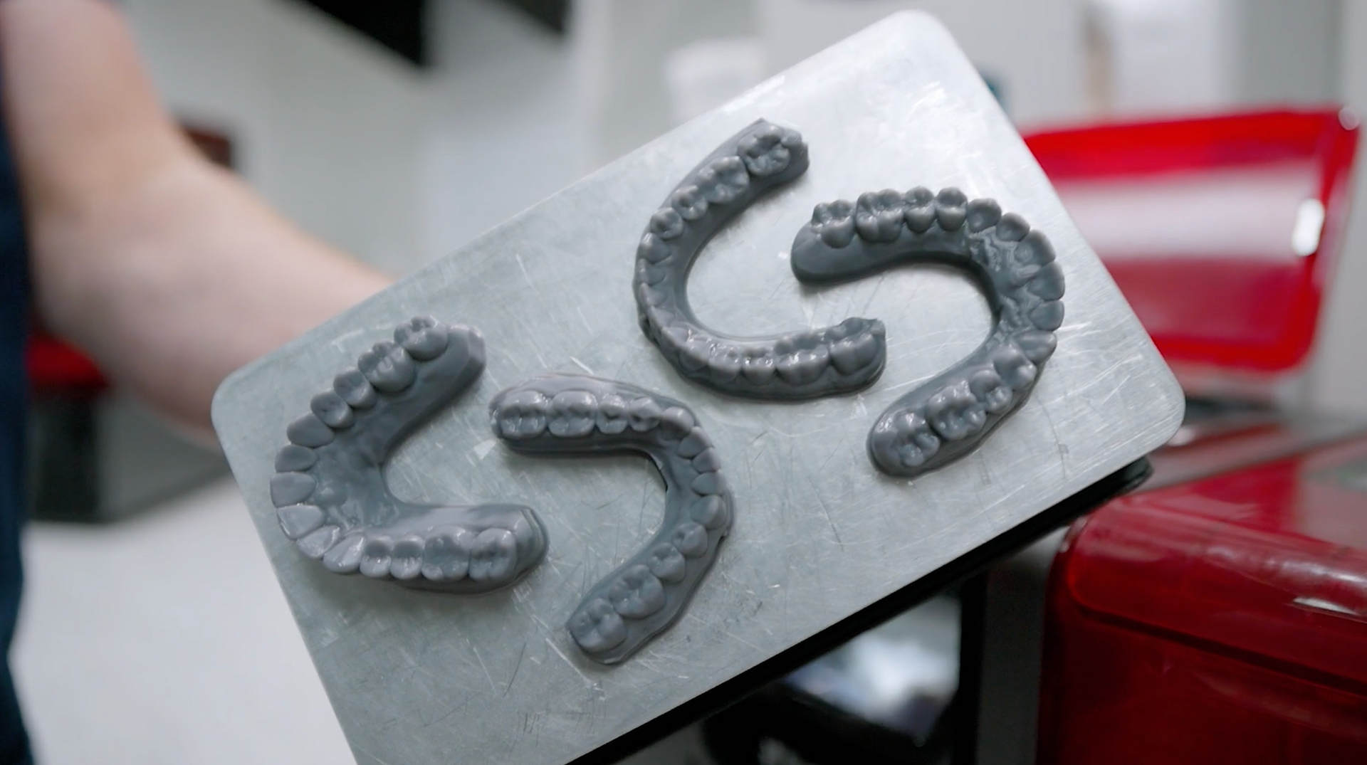 A person holding a metal tray with four gray, 3D-printed dental molds. Red object in the background.