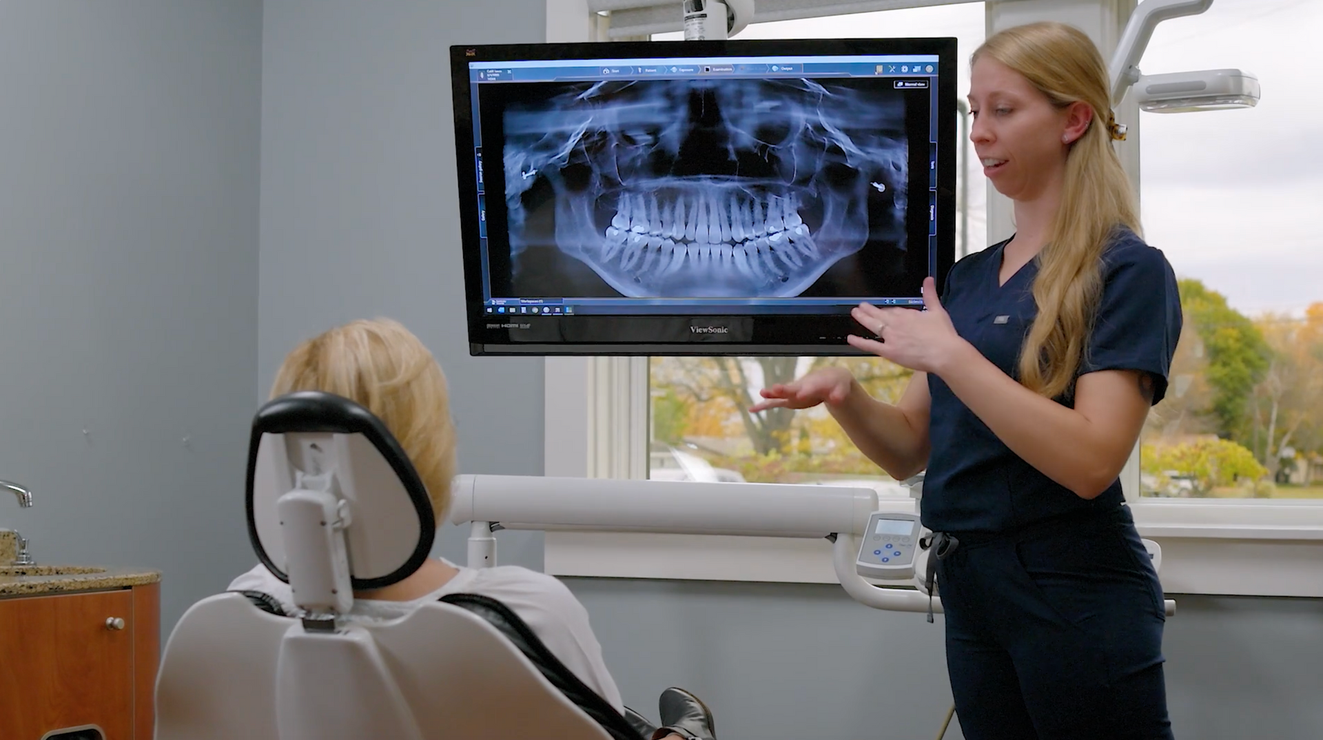 Woman smiling widely in a dental chair, facing a person out of frame, in a brightly lit office.