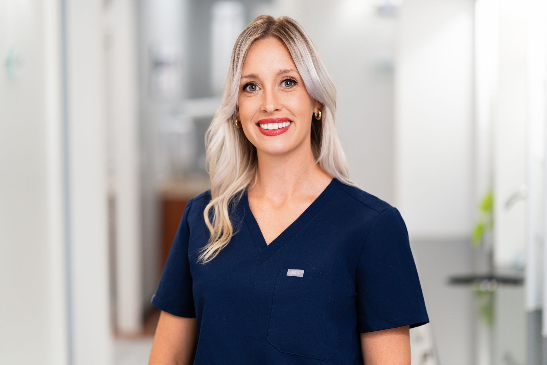 Woman with blonde hair, smiling, wearing navy scrubs, standing in a brightly lit hallway.