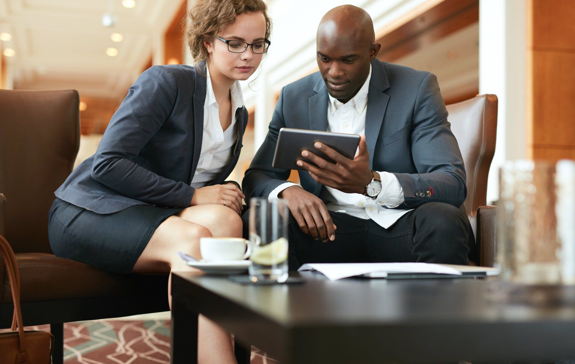 Two people in business attire sit closely, focused on a tablet screen in a modern office setting. A table with a glass of water and a cup is in the foreground.