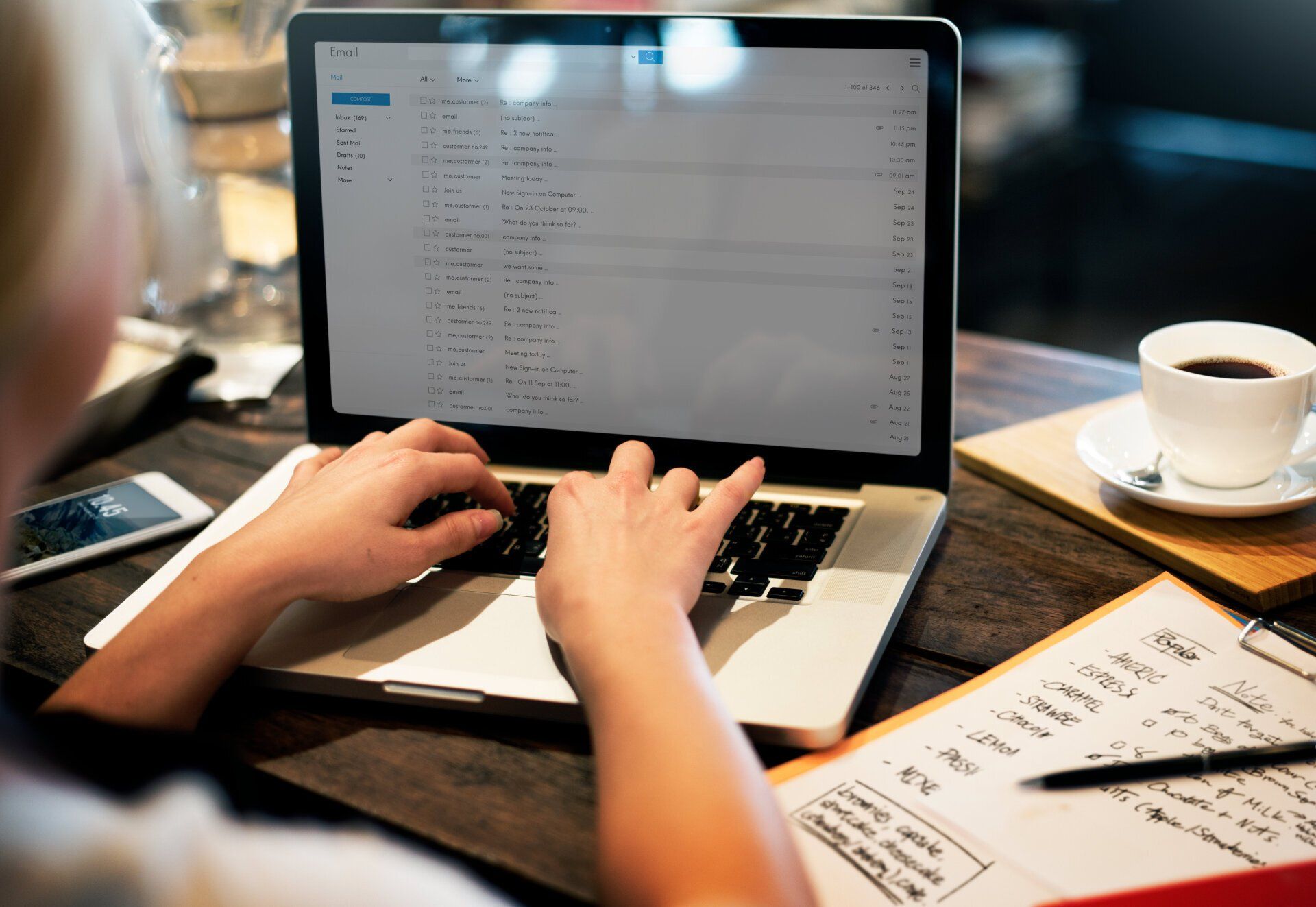 Person typing on a laptop displaying an email inbox, near a notebook with notes, a smartphone, and a cup of coffee on a table.