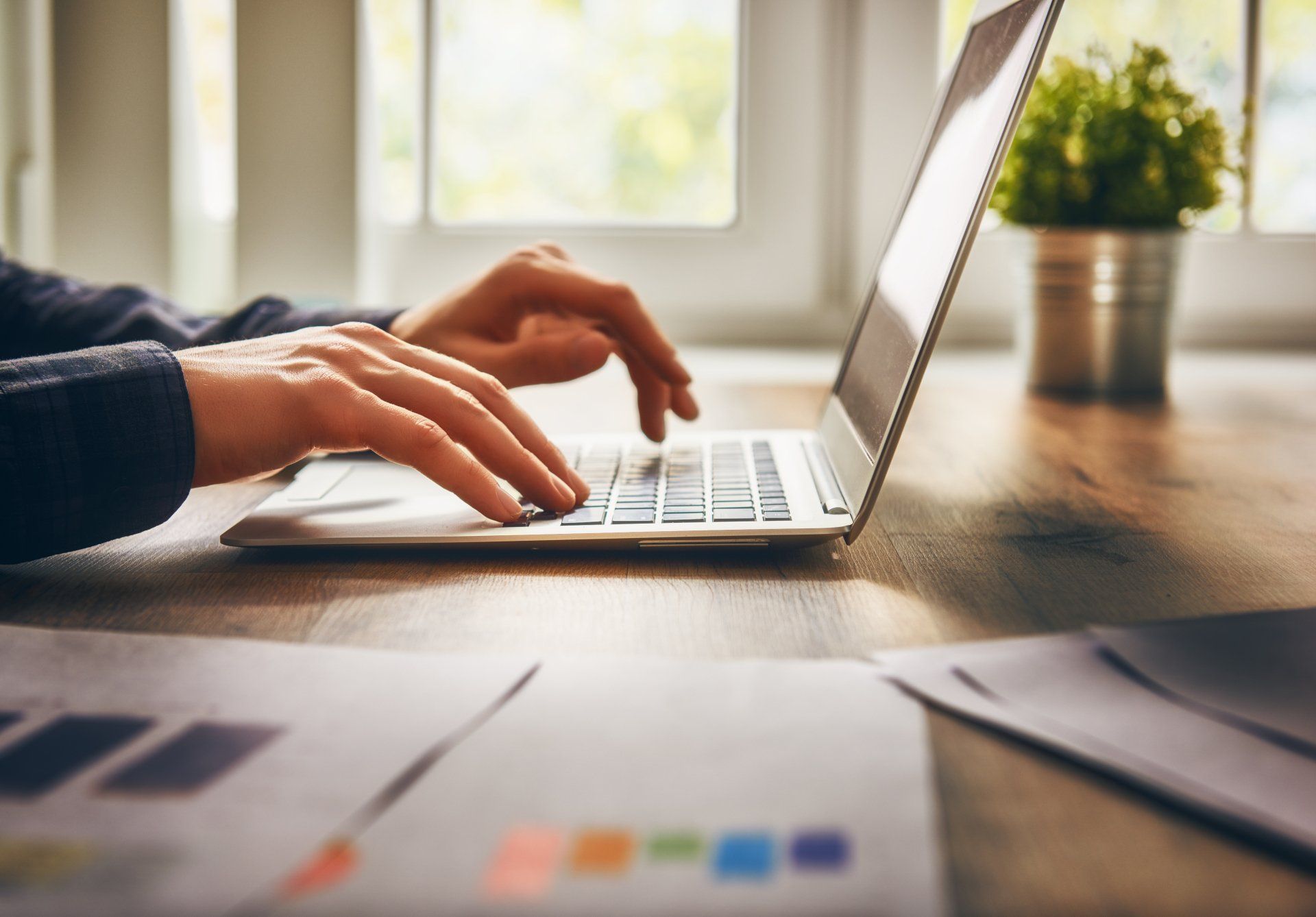 Hands typing on a laptop placed on a wooden desk, with papers in the foreground and a small plant by the window in the background.