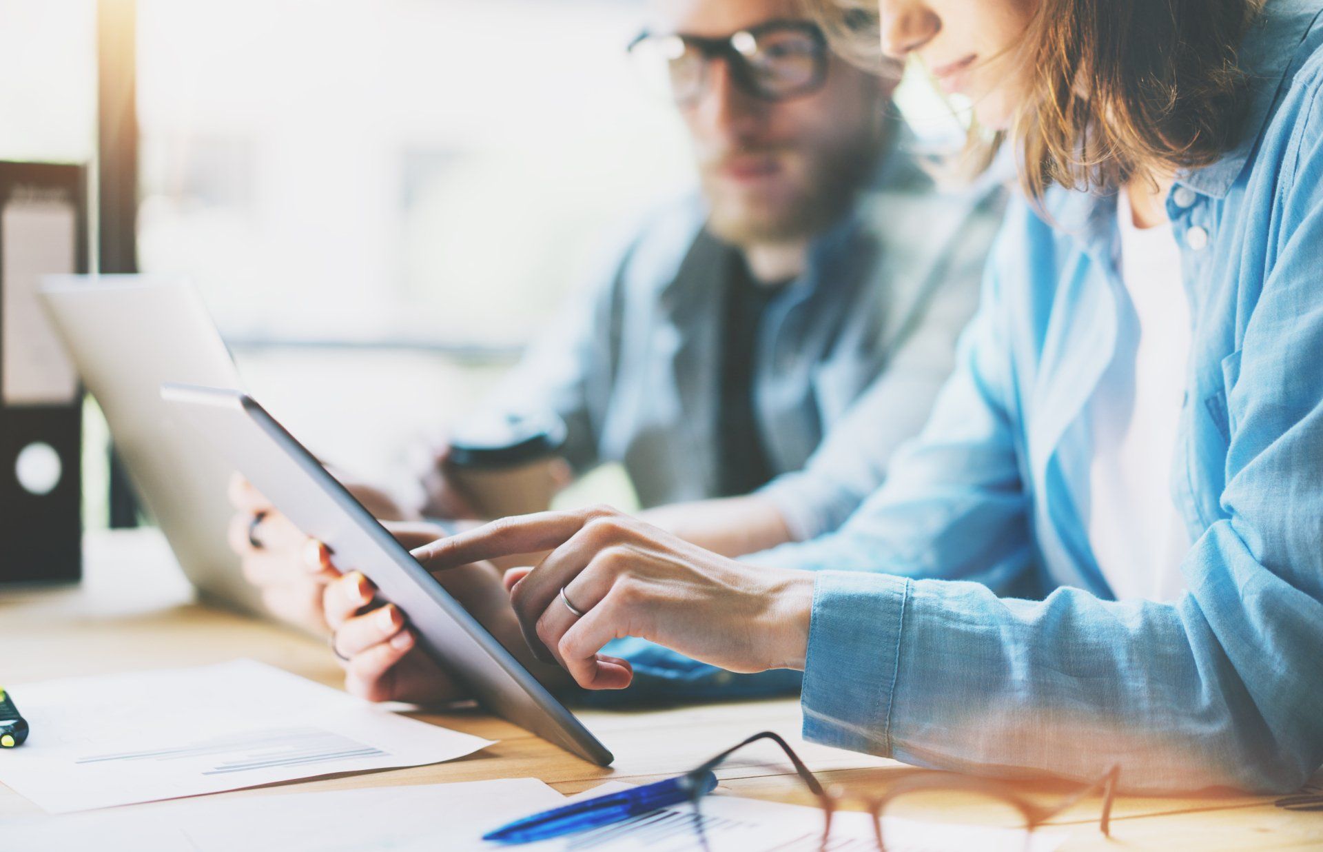 Two people sit at a table, focusing on a tablet. One is pointing at the screen. There are papers and pens on the table, and another laptop is visible.