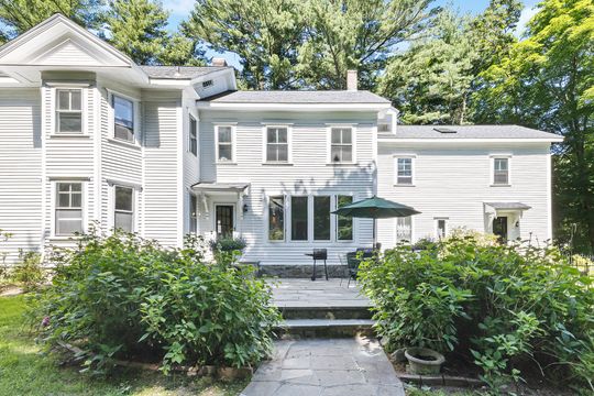 White two-story house with a front porch, steps, and green shrubs in bright sunlight