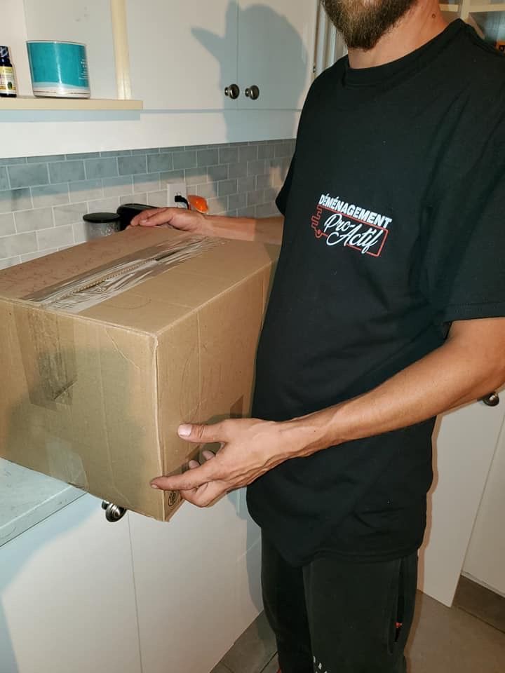 Un homme en chemise noire tient une boîte en carton dans une cuisine.