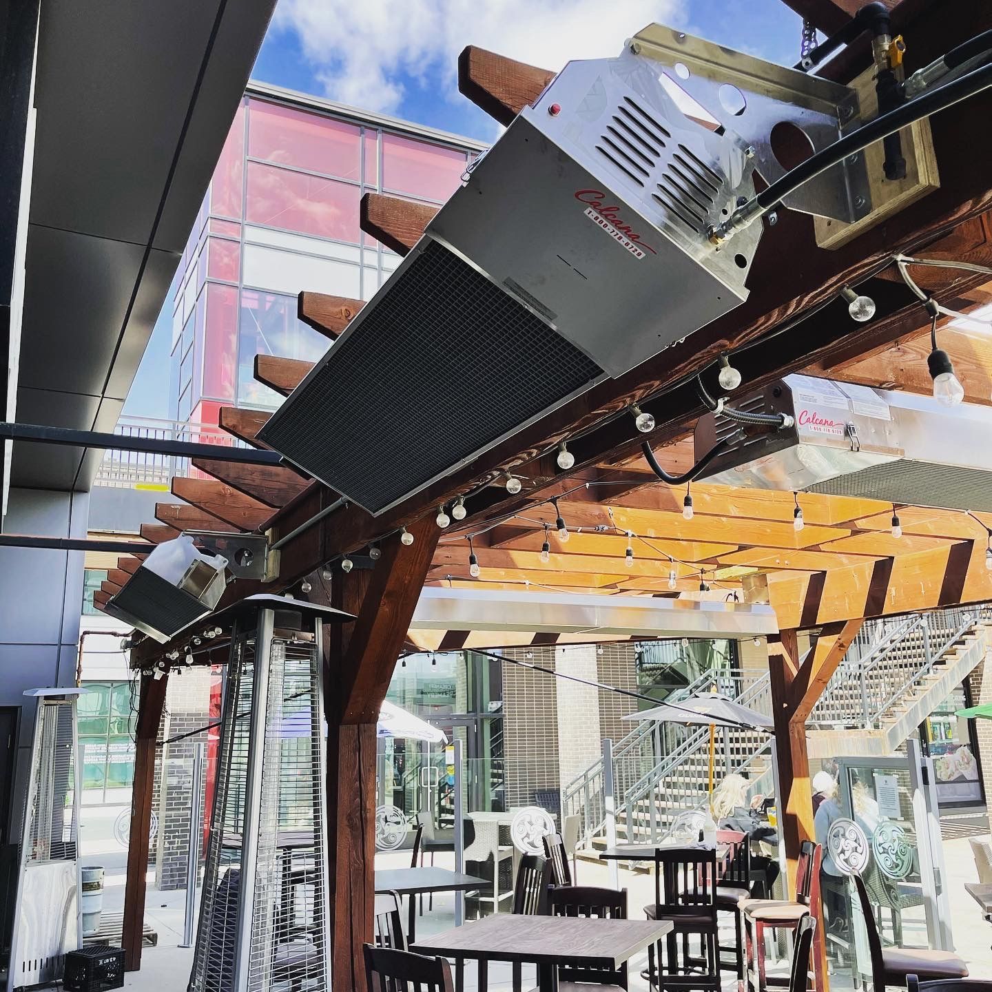 A restaurant with tables and chairs under a pergola with a heater hanging from the ceiling.